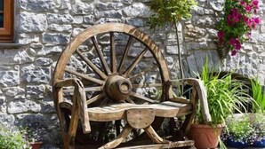 Un banc en bois rustique, doté d’une grande roue, placé contre un mur de pierres, avec des plantes et des fleurs en pot.