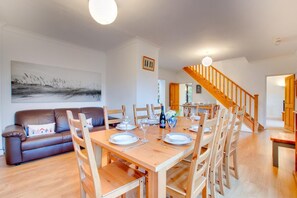 Un coin repas avec une table en bois dressée pour six personnes, entourée de chaises en bois. Un canapé en cuir marron est adossé au mur, et un escalier avec des rampes en bois est visible au fond.