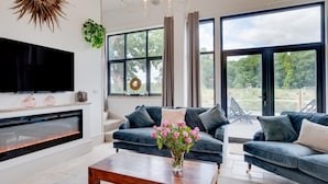 A modern living room with a fireplace, a television, and a large window with a view of greenery. - 5 Bed in Briston (oc-2305) (Briston)
