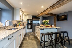 A modern kitchen with granite countertops, white cabinetry, a stainless steel refrigerator, and a central island with stools. - 5 Bed in Padstow (oc-p00595) (Padstow)
