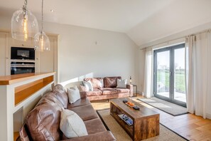A modern living room with a leather sofa, wooden coffee table, and a kitchen area with an oven. - 3 Bed in North Sunderland (oc-t33656) (North Sunderland)