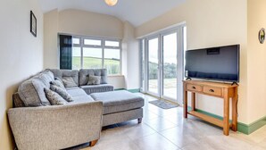 A modern living room with a grey sectional sofa, a flat-screen TV on a wooden stand, and large sliding glass doors leading to a balcony. - 3 Bed in Llanon (oc-porth) (Llan-non)