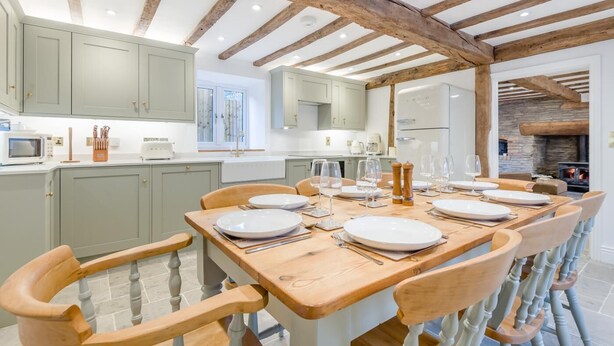 A modern kitchen with light green cabinets, wooden beams, and a dining area with a wooden table set for six. - 4 Bed in Peterchurch (oc-s34019) (Peterchurch)