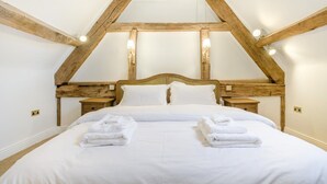 A bedroom with a high ceiling, exposed wooden beams, a large bed with white linens, two bedside tables, and wall-mounted lights. - 4 Bed in Peterchurch (oc-s34019) (Peterchurch)
