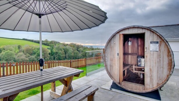 An outdoor wooden sauna with a bench and an umbrella, surrounded by a wooden fence and a view of a green landscape. - 2 Bed in Llanon (oc-porth1) (Llan-non)