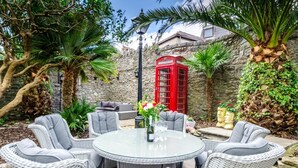 A round glass table with wicker chairs, a red telephone booth, and lush greenery in a courtyard setting. - 4 Bed in Tenby (oc-pw9264) (Tenby)