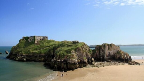 A coastal landscape with a grassy hill and a building on top, overlooking a sandy beach and calm sea. - 4 Bed in Tenby (oc-pw9264) (Tenby)