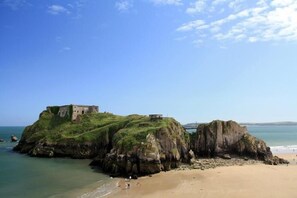A coastal landscape with a grassy hill and a building on top, overlooking a sandy beach and calm sea. - 4 Bed in Tenby (oc-pw9264) (Tenby)