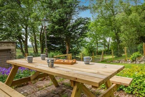 Ein Holztisch für ein Picknick mit gestreiften Tassen und einem Korb voller Muffins, gelegen in einem Garten mit einer Laterne und einer Steinmauer.