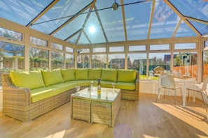 A conservatory with a curved green sofa, a glass coffee table, and a dining area with chairs and a table. - 3 Bed in Langham (oc-1828) (Langham)