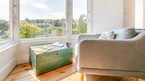 Una habitación con un sofá gris, un baúl verde y vistas a espacios verdes y edificios a través de ventanas amplias.