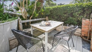 Una terraza de madera con una mesa y dos sillas, rodeada de abundante vegetación y un muro de ladrillo.