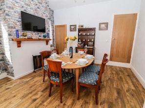 A dining area with a brick wall, a flat-screen TV, a wooden dining table set for six, and a bookshelf with decorative items.