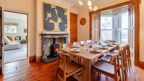Une salle à manger avec une cheminée, une table à manger en bois dressée pour six personnes et une vue sur l’extérieur grâce à de grandes fenêtres.
