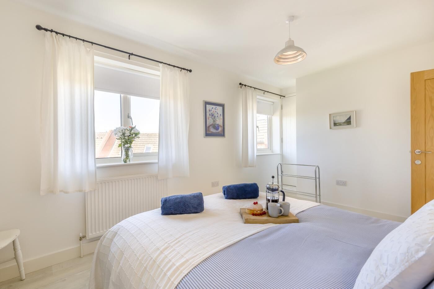 A bedroom with a bed, a window with curtains, a radiator, a bedside table, a hanging light fixture, and a door.