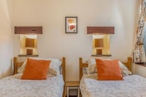 Ein Zimmer mit zwei Betten, jedes mit einem hölzernen Kopfende und einem orangefarbenen Kissen. Über jedem Bett befindet sich eine Wandlampe und zwischen den Betten hängt ein gerahmtes Bild.