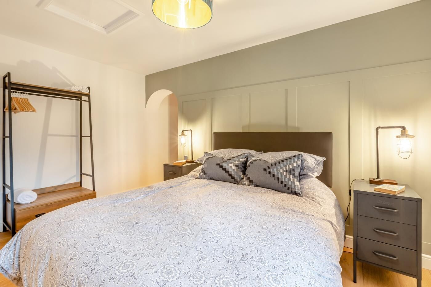 A bedroom with a large bed, a bedside table with a lamp, a wardrobe with a mirror, and a hanging rack.