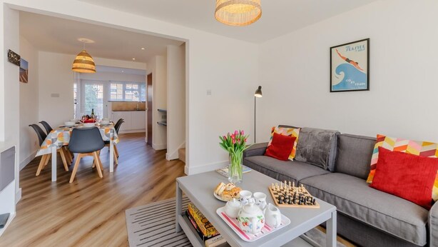 Una sala de estar moderna con un sofá gris, una zona de comedor con una mesa dispuesta para dos personas y una cocina visible al fondo.