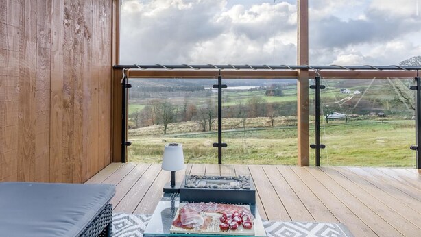 Une terrasse en bois dotée d’une balustrade en verre, offrant une vue sur un paysage rural.