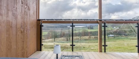 Une terrasse en bois dotée d’une balustrade en verre, offrant une vue sur un paysage rural.