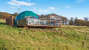 Une serre en verre, dotée d’une structure métallique et recouverte en partie d’une bâche verte, située sur une colline herbeuse.