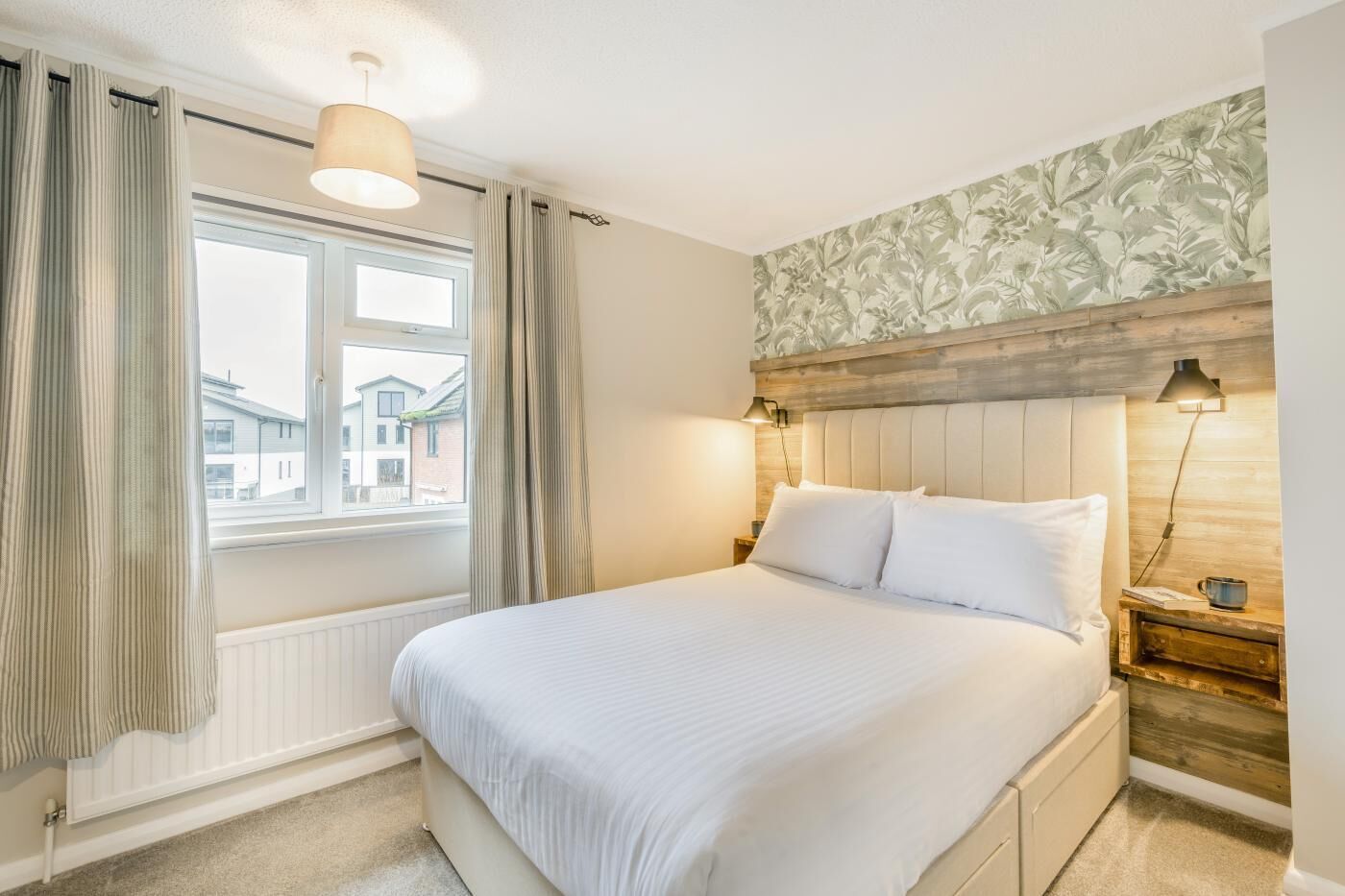 Ein Schlafzimmer mit einem großen Bett, einem Fenster mit Vorhängen, einem Holz-Kopfteil und einem Nachttisch mit Lampe.