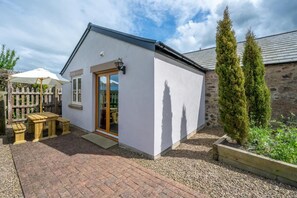 A small, single-story building with a white exterior, a wooden door, and a small patio area with a table and benches. - 2 Bed in Milfield (oc-nb1982) (Milfield)