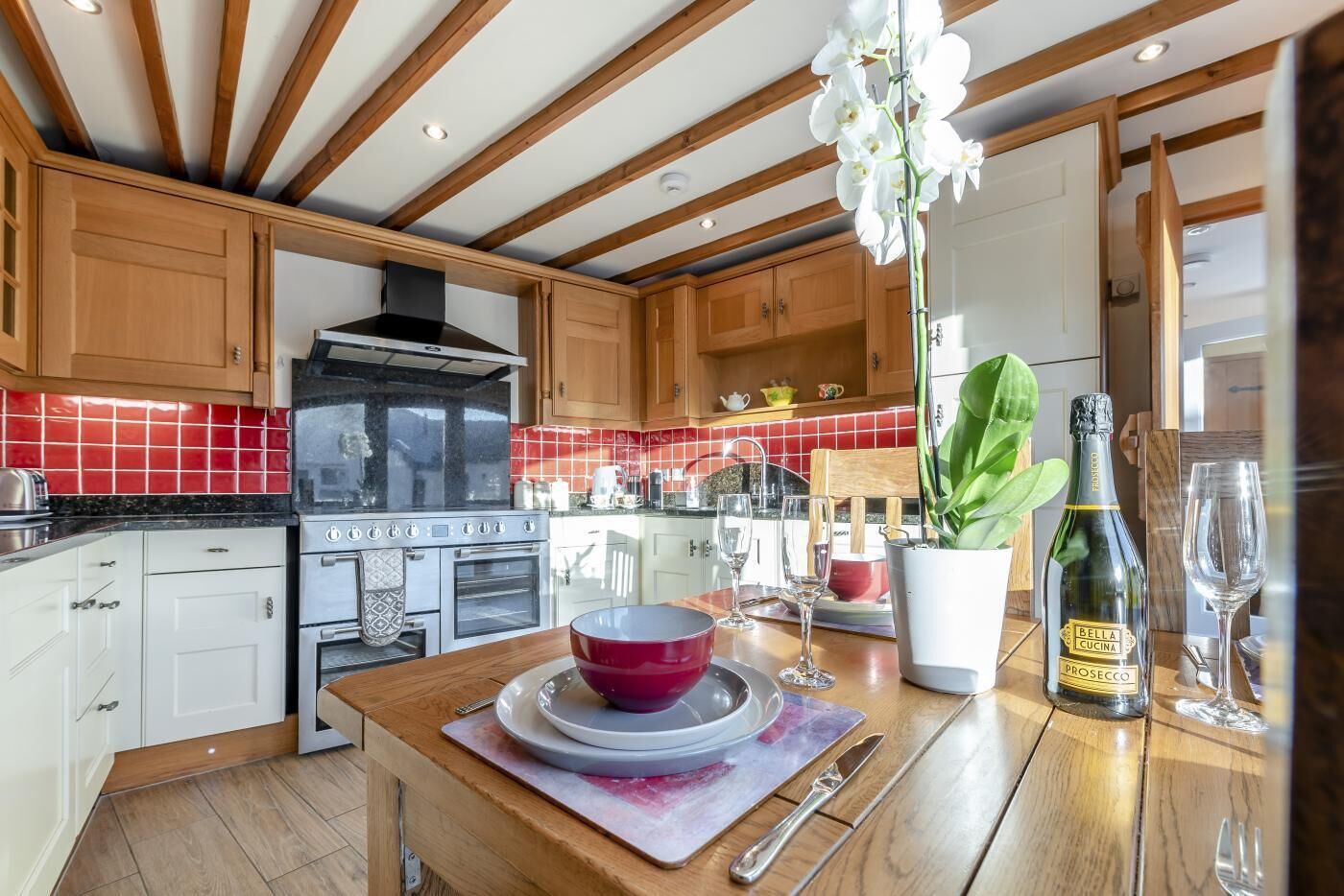 A modern kitchen with wooden beams, red-tiled backsplash, stainless steel appliances, and a dining area with a table set for two.