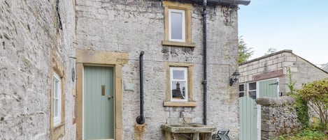 A stone building with two visible doors and windows, a small outdoor seating area, and a garden.