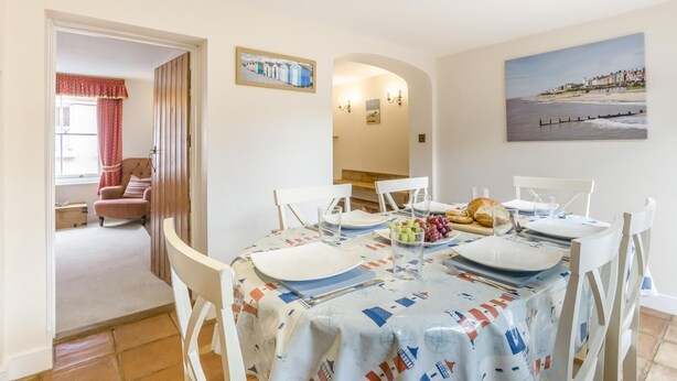 A dining area with a table set for four, a chair, and a view of the beach through a window. - 3 Bed in Southwold (oc-stan) (Southwold)