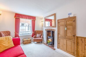A cozy living room with a fireplace, a red sofa, a wooden cabinet, and a window with red curtains. - 3 Bed in Southwold (oc-stan) (Southwold)