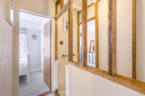 Un couloir avec des poutres en bois, une porte blanche et une vue sur une pièce où l’on aperçoit un lit et un radiateur.