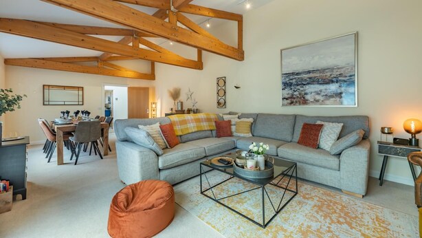 A spacious living area with a large grey sofa, wooden beams on the ceiling, a dining table with chairs, and a painting on the wall.