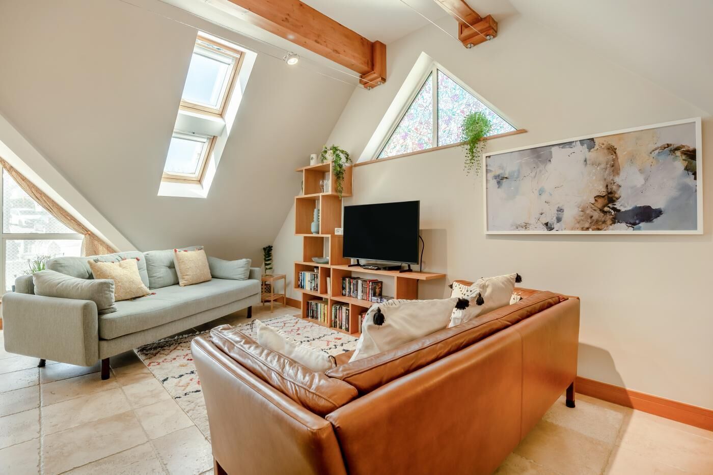 A modern loft-style living room with a high ceiling, exposed wooden beams, a large window, a built-in TV shelf, and a large abstract painting.