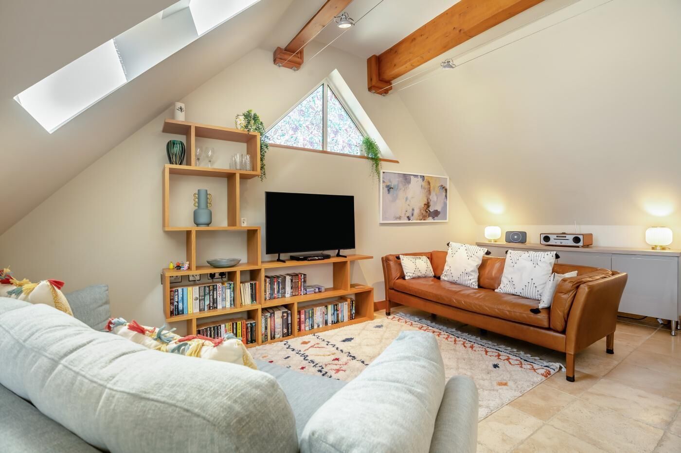 Ein gemütliches Wohnzimmer im Loft-Stil mit schrägem Dach, einem Fernseher, einem Bücherregal, einer Couch und einer kleinen Küchenzeile mit Radio und Lampe.