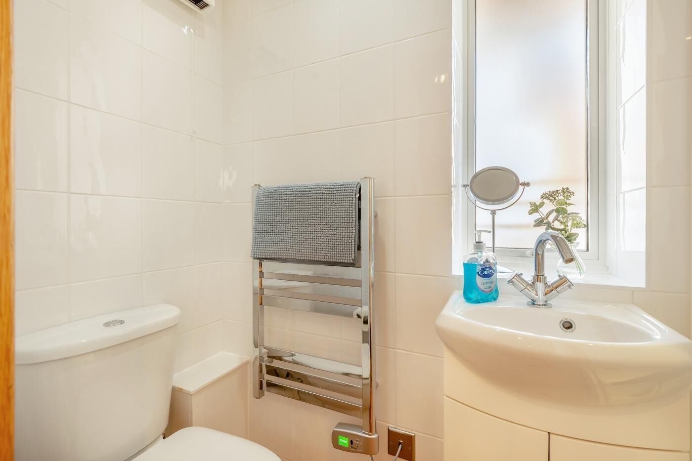 A bathroom with white tiles, a towel rack, a toilet, a sink with a mirror, and a window.