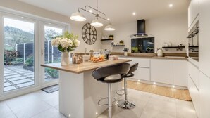 A modern kitchen with a central island, bar stools, and a large window overlooking a garden. - 3 Bed in Brithdir (oc-c33971) (Brithdir)