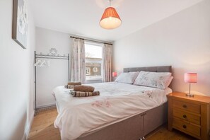 Een slaapkamer met een bed, nachtkastje, lamp, raam met gordijnen en een uitzicht op gebouwen.
