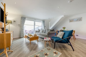Un salon moderne avec un canapé, une table basse et un fauteuil bleu. Un escalier mène à un niveau supérieur, et on y trouve un coin repas avec une table et des chaises.
