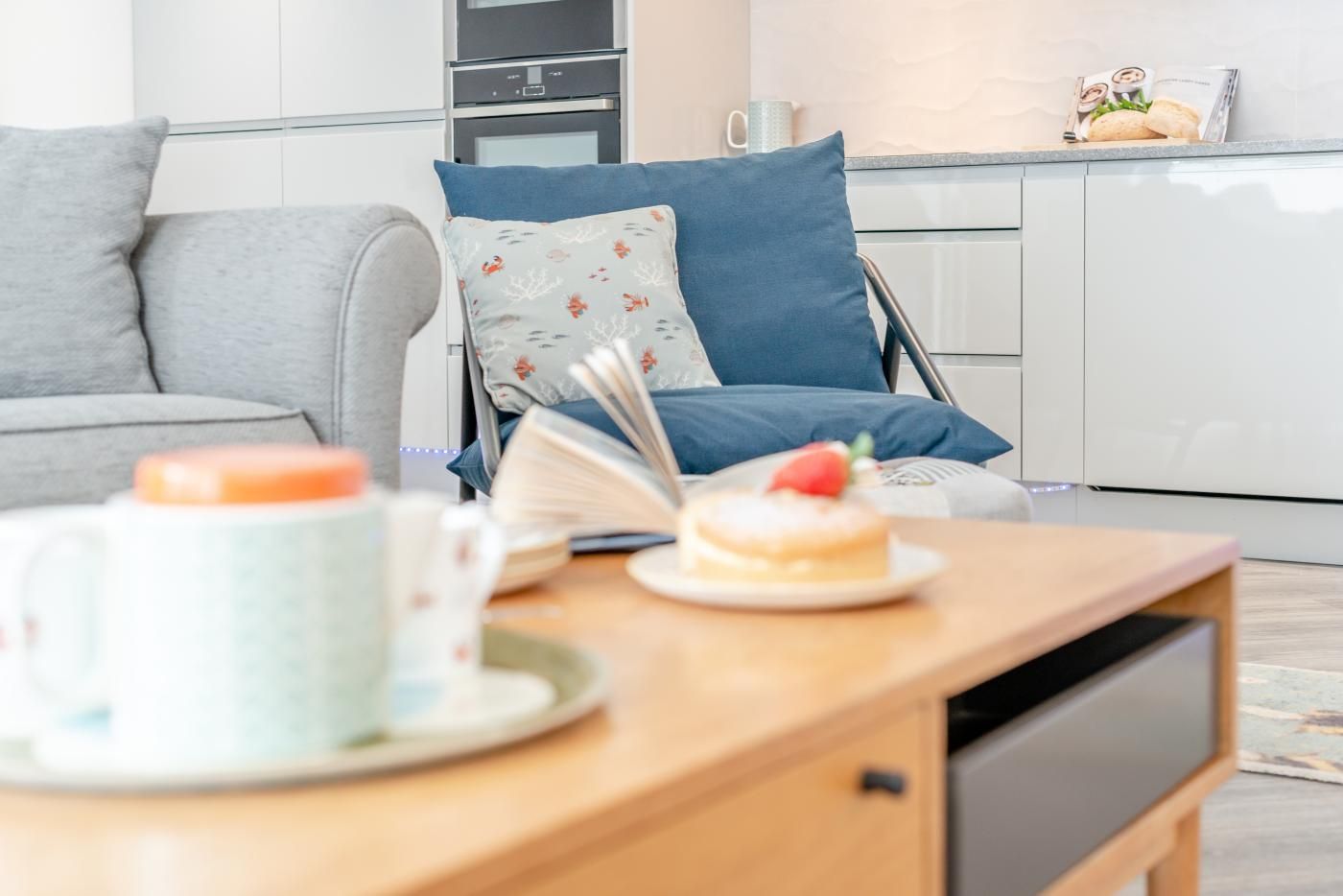 Ett modernt kök med vita skåp, en grå soffa och ett träbord med en bok, en mugg och en tallrik med en efterrätt på.