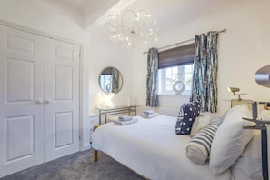 Ein Schlafzimmer mit einem Bett, Nachttischen, einem Spiegel, einer Chandelüster, Vorhängen und einem Fenster.