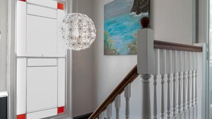 Un escalier avec des balustrades blanches et une main courante en bois, menant à un palier où est accrochée une peinture représentant un paysage.
