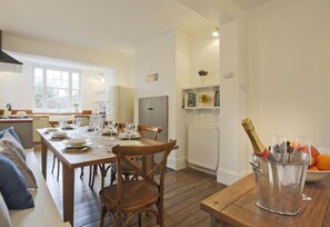 Un comedor con una mesa de madera dispuesta para seis comensales, una cocina al fondo y una botella de champán sobre una mesita.