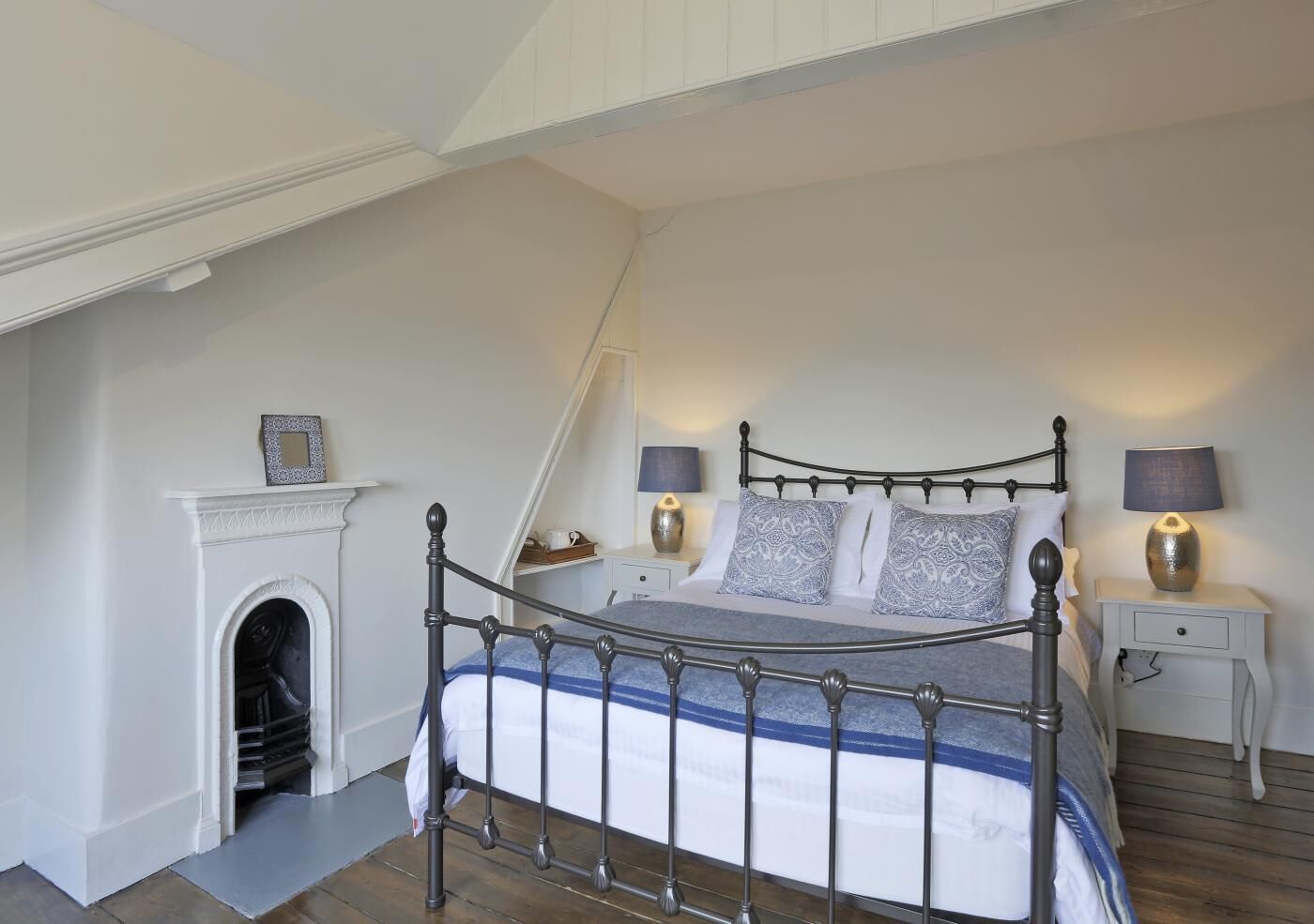 A bedroom with a metal bed frame, a fireplace, two bedside tables with lamps, and a staircase.