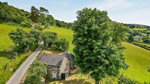 A stone house with a paved driveway, surrounded by lush green fields and trees. - 1 Bed in Llangower (oc-t34142) (Llangower)