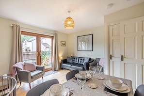 A dining area with a table set for four, a sofa, and a view of a house through a glass door. - 2 Bed in Nr. Newby Bridge (oc-ld168) (Backbarrow)
