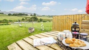 A picnic table with a book, glasses, mugs, and croissants, overlooking a grassy field with sheep and a wooden fence. - 3 Bed in Bala (oc-bow26) (Parc)