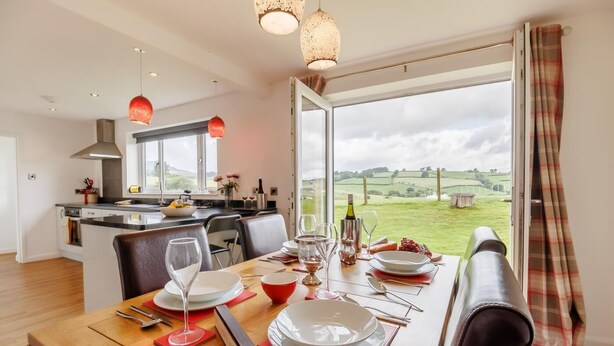 A dining area with a table set for two, a kitchen in the background, and a view of a green landscape through a large window. - 3 Bed in Bala (oc-bow26) (Parc)
