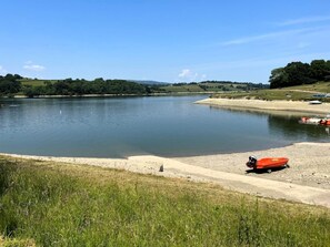 Ein ruhiger See mit sandigem Ufer, umgeben von Grünflächen und Hügeln.