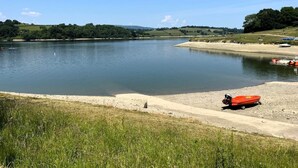 Un lago tranquilo con orilla arenosa, rodeado de vegetación y colinas.